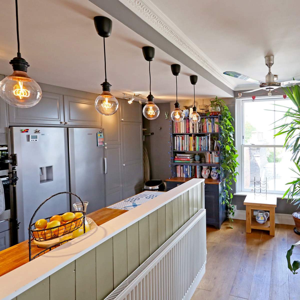 A bright, contemporary kitchen area featuring a curved countertop, hanging pendant lights, a bookshelf, and large windows with indoor plants.
