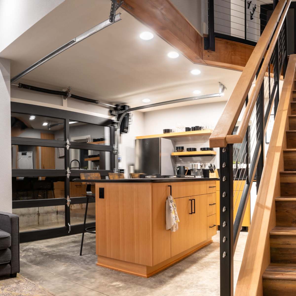 A modern interior with a staircase leading to an upper level, a gray couch on the left, and a kitchen area with wooden cabinetry and stainless steel appliances.