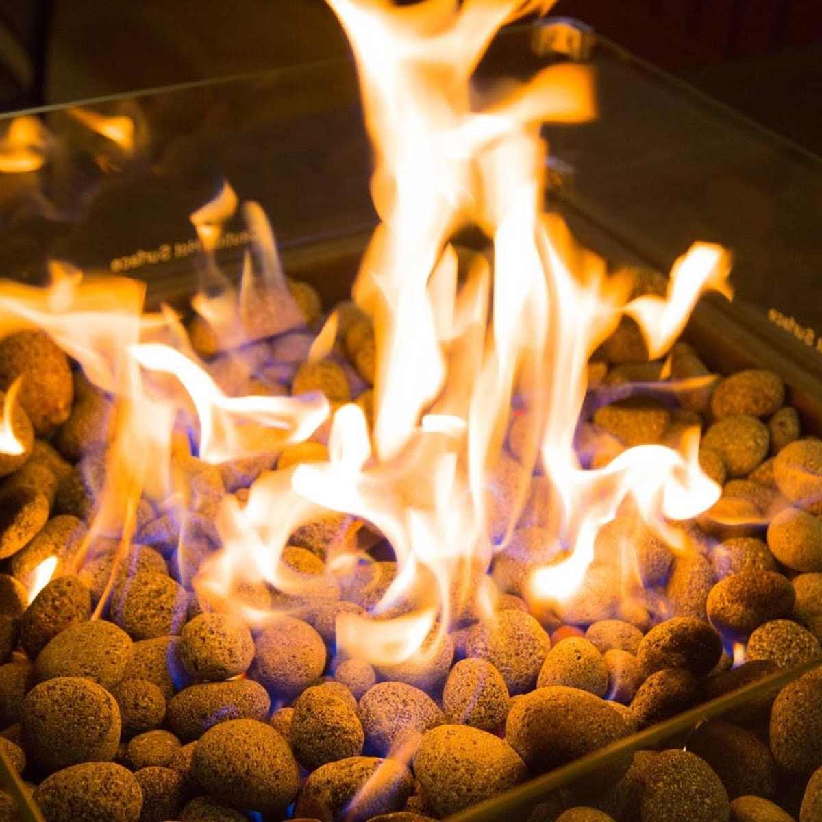 A fire burns brightly among smooth stones in a glass-enclosed fire pit.