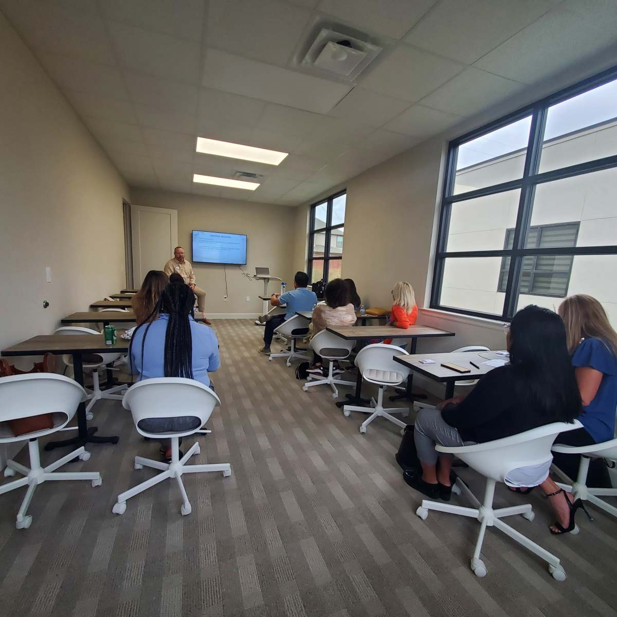 A group of people is seated in a modern classroom, attentively facing a presenter and a large screen displaying information.