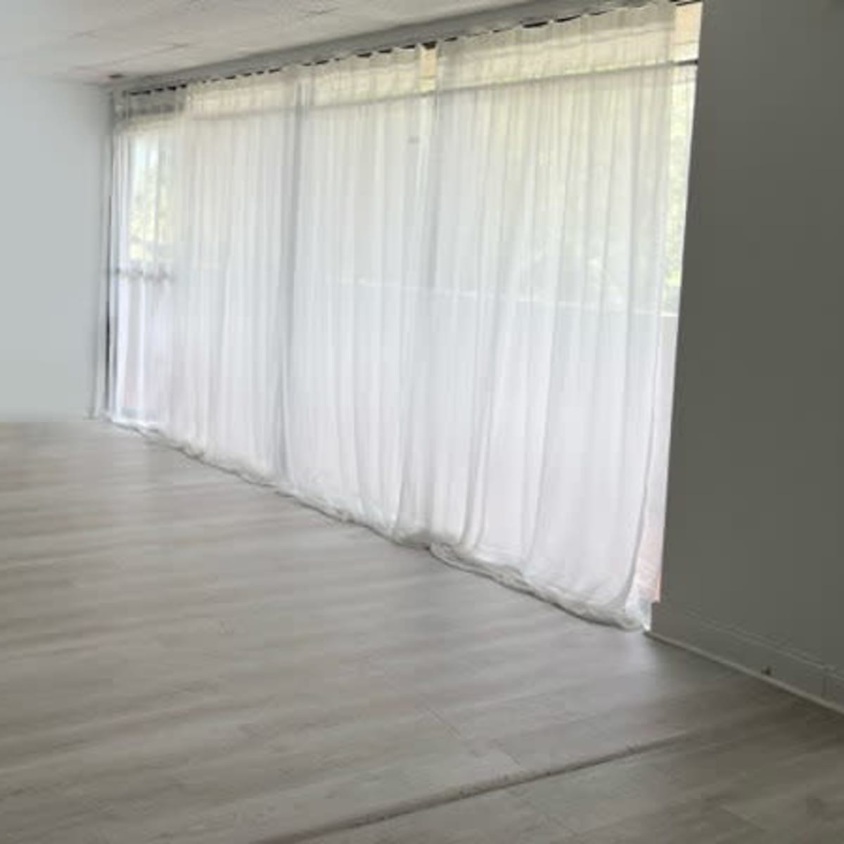 A spacious room with light wooden flooring and sheer curtains covering a large window.