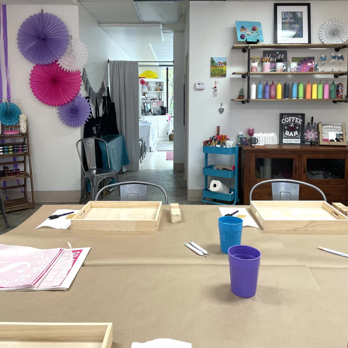 A craft studio with wooden trays on a table covered in brown paper, and shelves filled with colorful paint and supplies in the background.