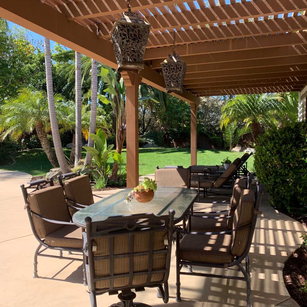 A covered outdoor dining area features a glass table surrounded by chairs, with palm trees and a grassy lawn in the background.