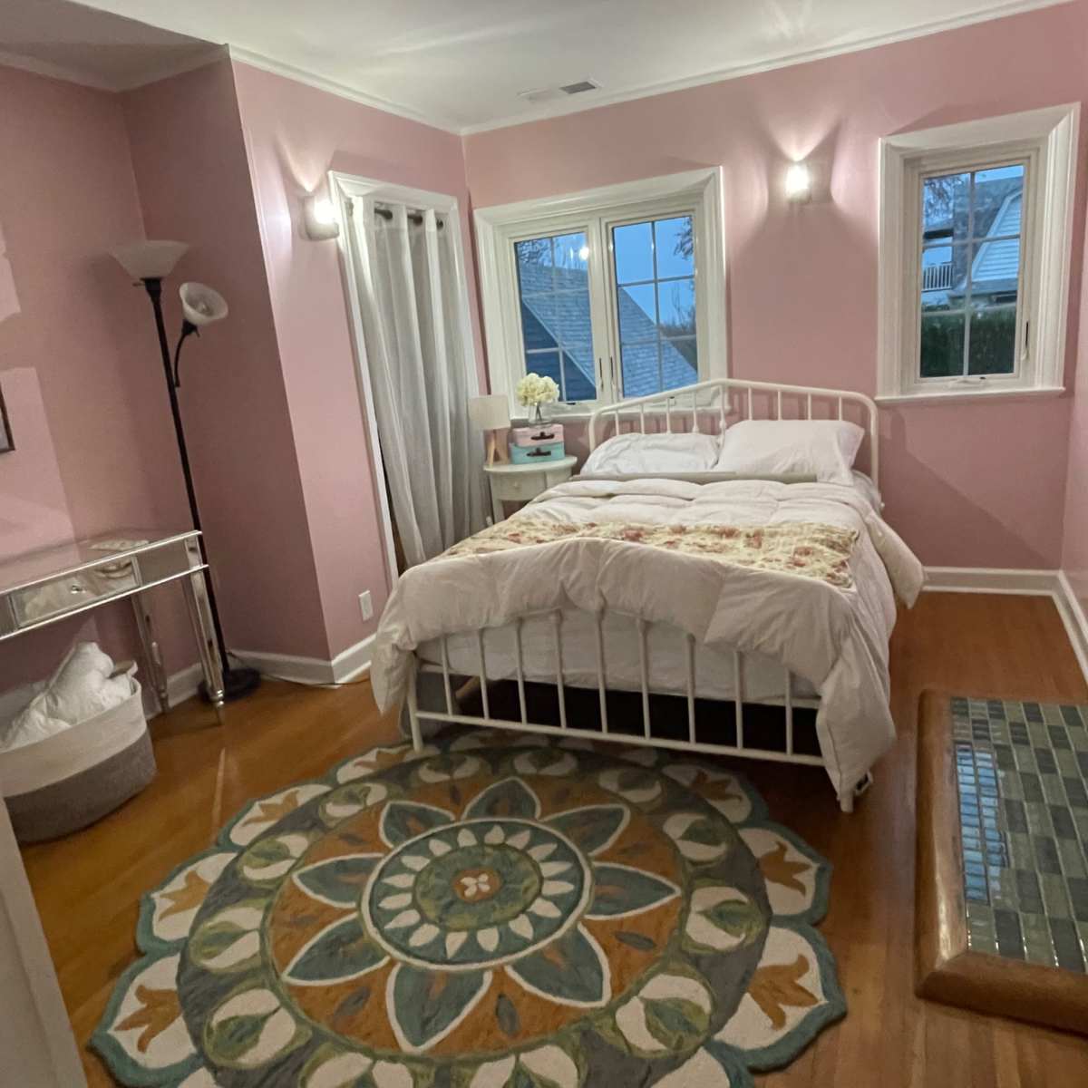 A bright pink bedroom featuring a bed with white bedding, a circular patterned rug on the hardwood floor, and a fireplace with green tiles.