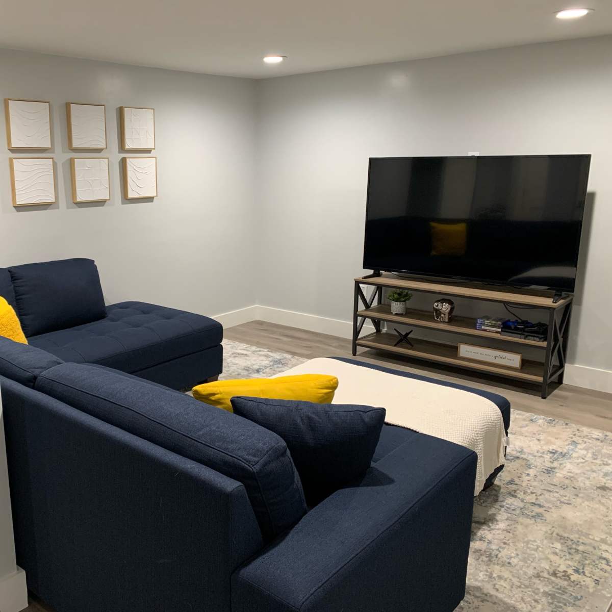 A modern basement living room features a dark blue sectional sofa, a wooden TV stand with a television, and framed artworks on the wall.