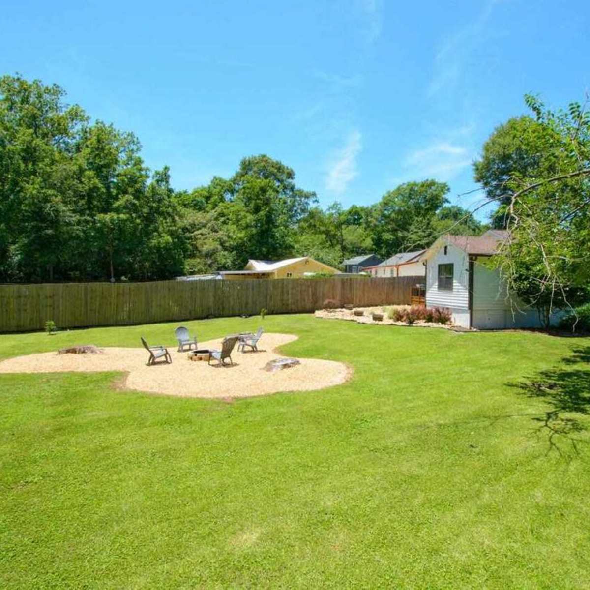 A spacious backyard features a circular gravel area with chairs arranged around a fire pit, surrounded by a green lawn and trees.