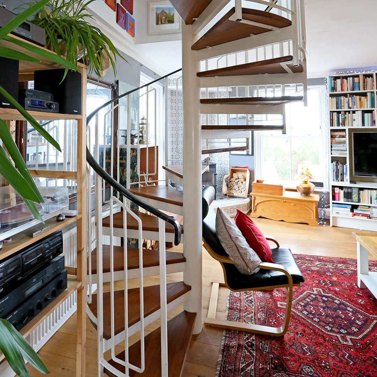 A spiral staircase with wooden steps and a glass railing is situated in a well-furnished living area featuring bookshelves and decorative elements.