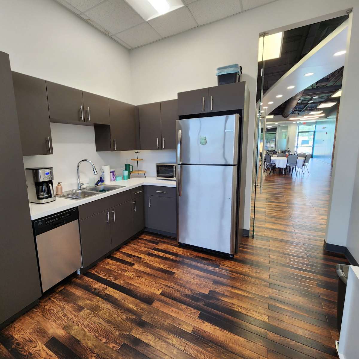 A modern kitchen with dark cabinets, a stainless steel refrigerator, and hardwood flooring, leading into an open area beyond.