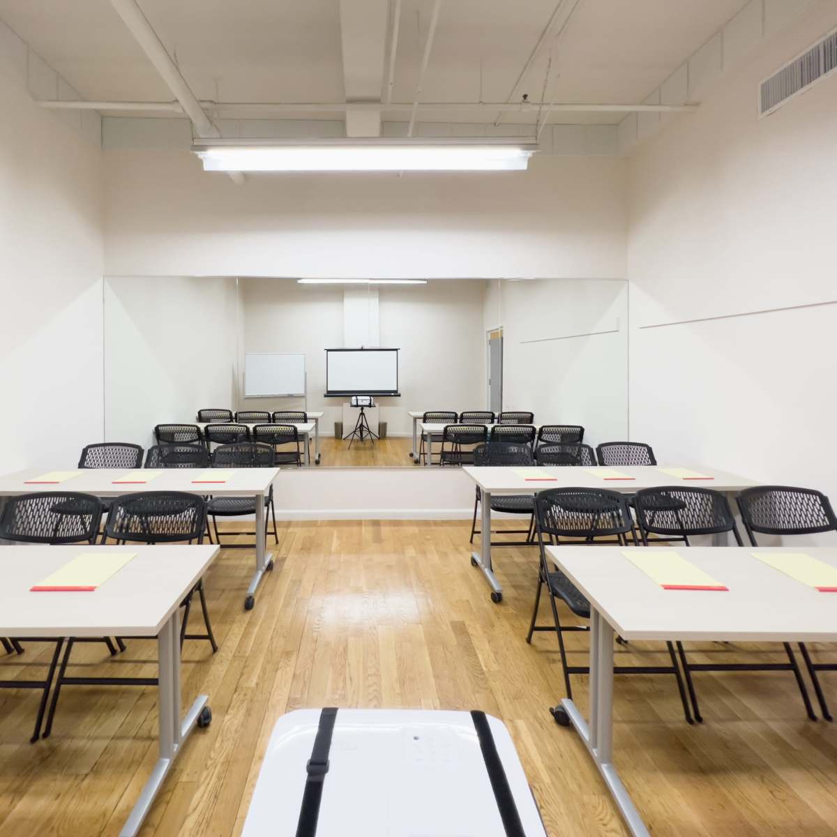 A classroom setting features multiple tables with chairs arranged facing a screen and a large mirror on one wall.