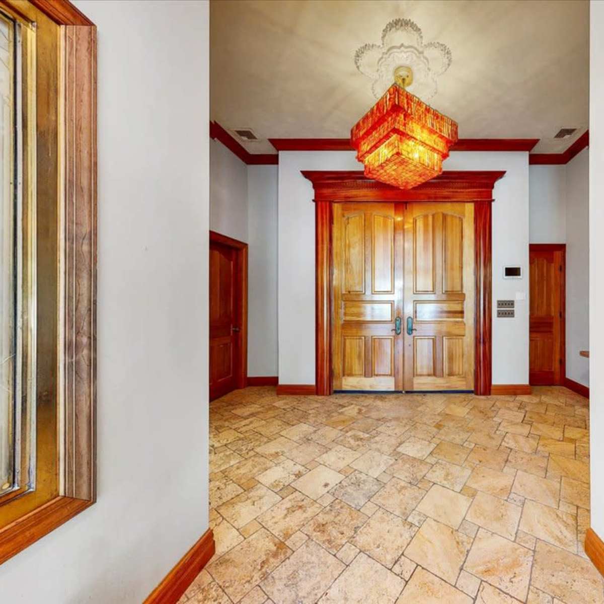 A hallway leading to large wooden double doors, with a chandelier hanging above and tiled flooring.