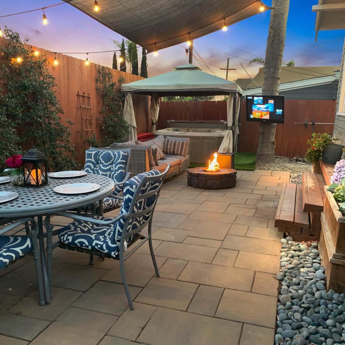 A patio area with a circular fire pit, dining table set, comfortable seating, and ambient lights strung overhead.