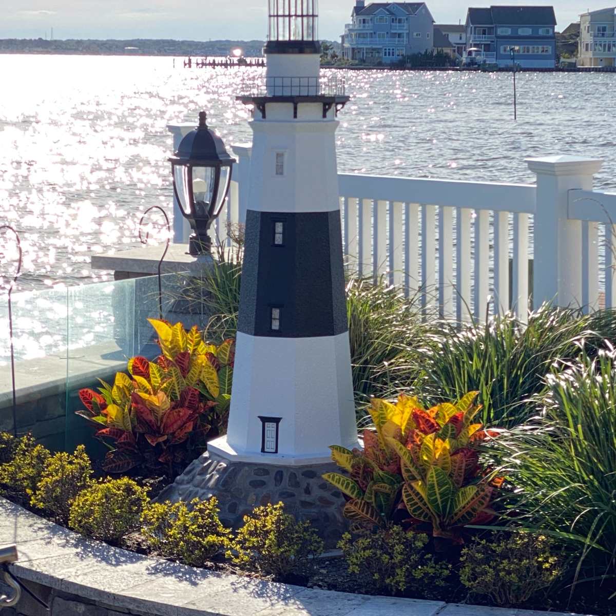 A small lighthouse sculpture surrounded by colorful plants sits on a stone terrace overlooking a shimmering body of water.