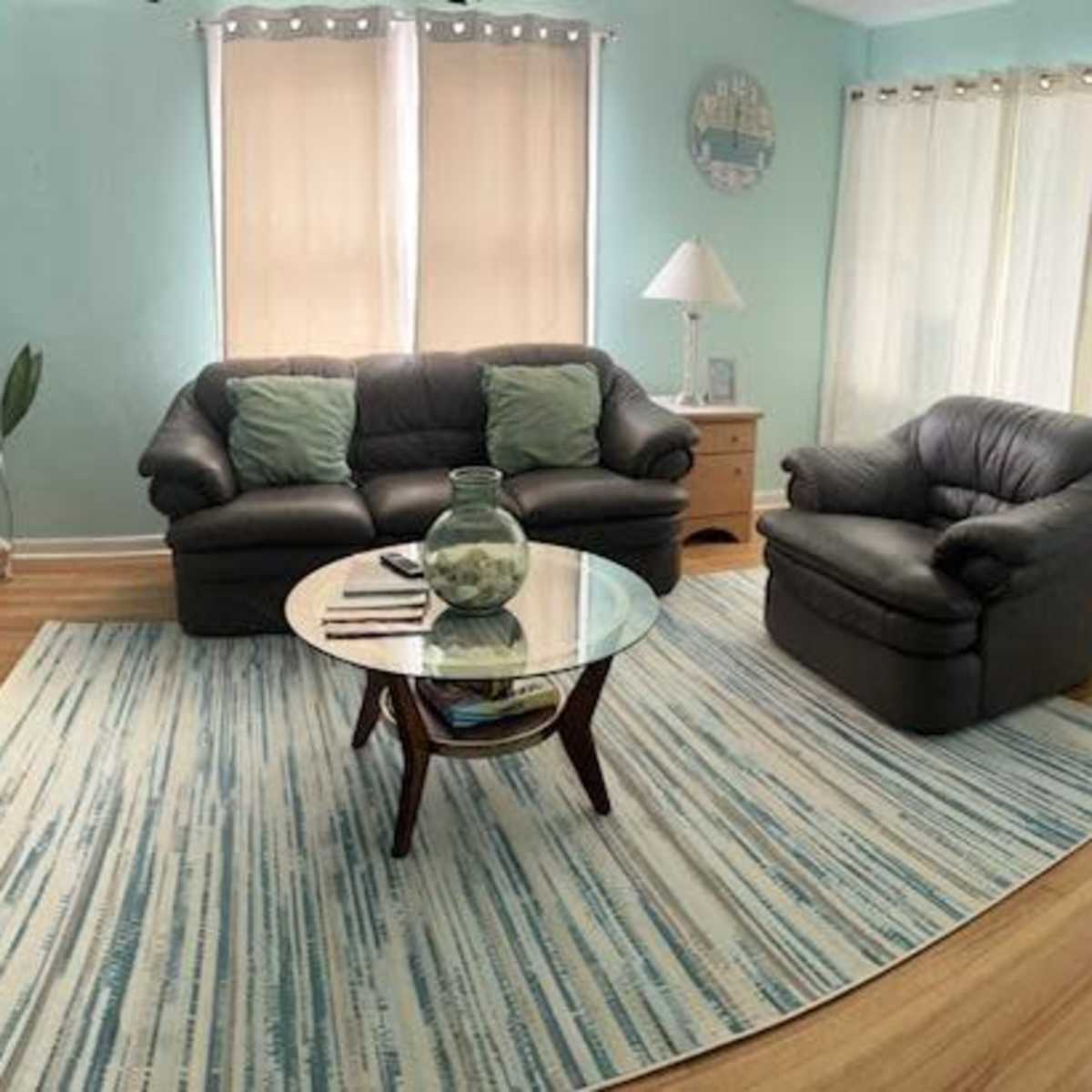 A light blue living room with a black leather sofa, a matching armchair, a round glass coffee table, and large windows dressed with sheer curtains.