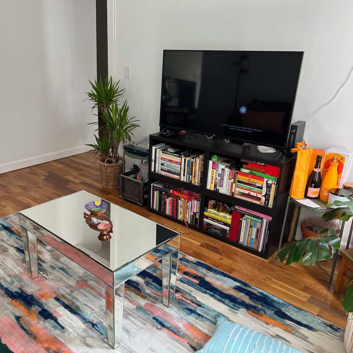 A living room features a mirrored coffee table, a dark blue sofa, a bookshelf filled with books, and a large television mounted on the wall.