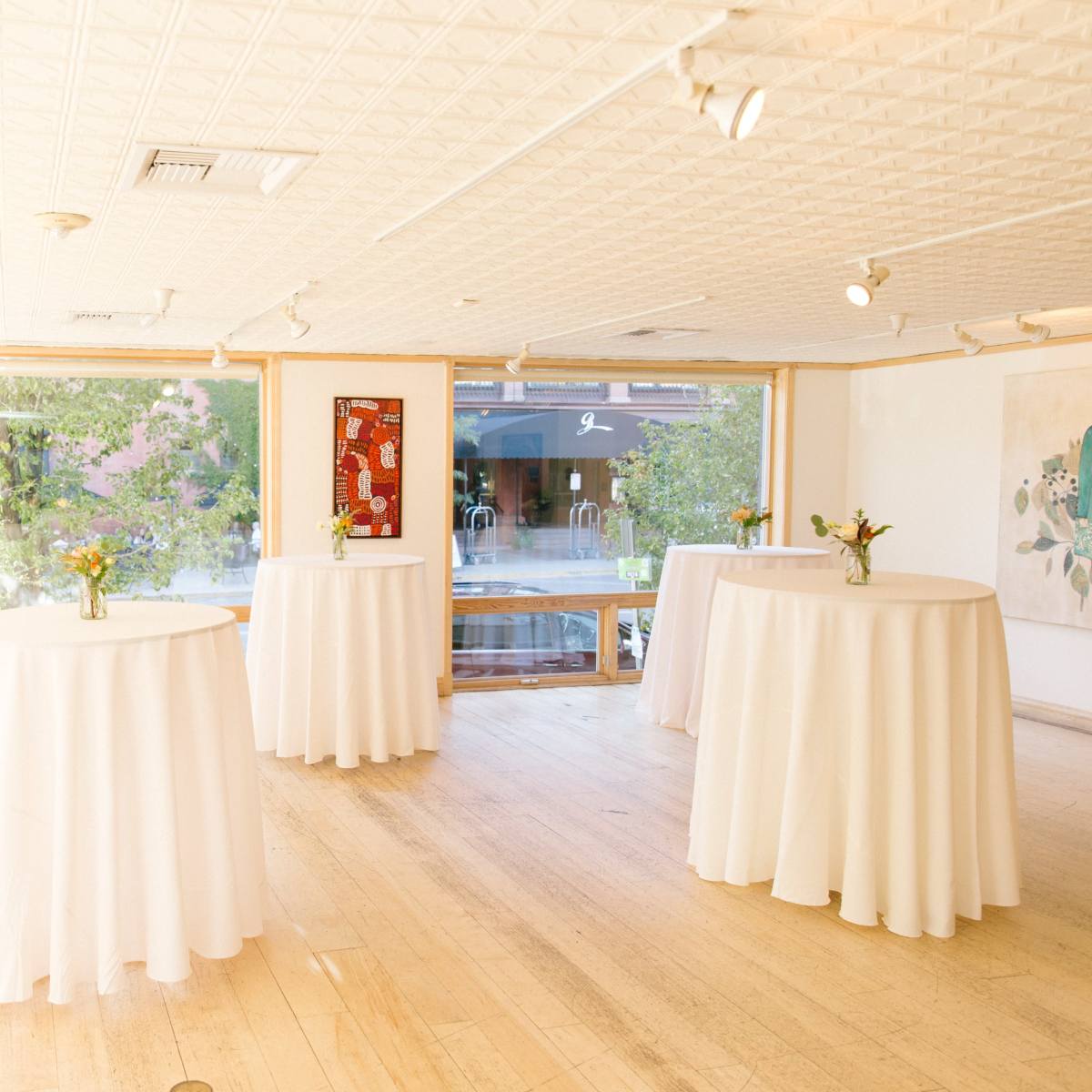 A bright gallery space featuring several round tables dressed in white tablecloths, with windows overlooking a tree-lined street.