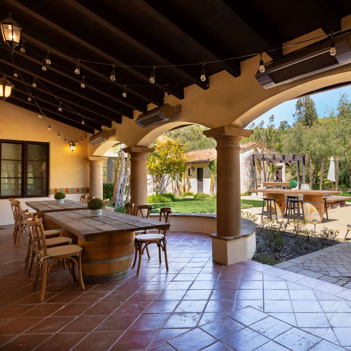 An outdoor patio area featuring a long wooden table, chairs, and lush greenery in the background.