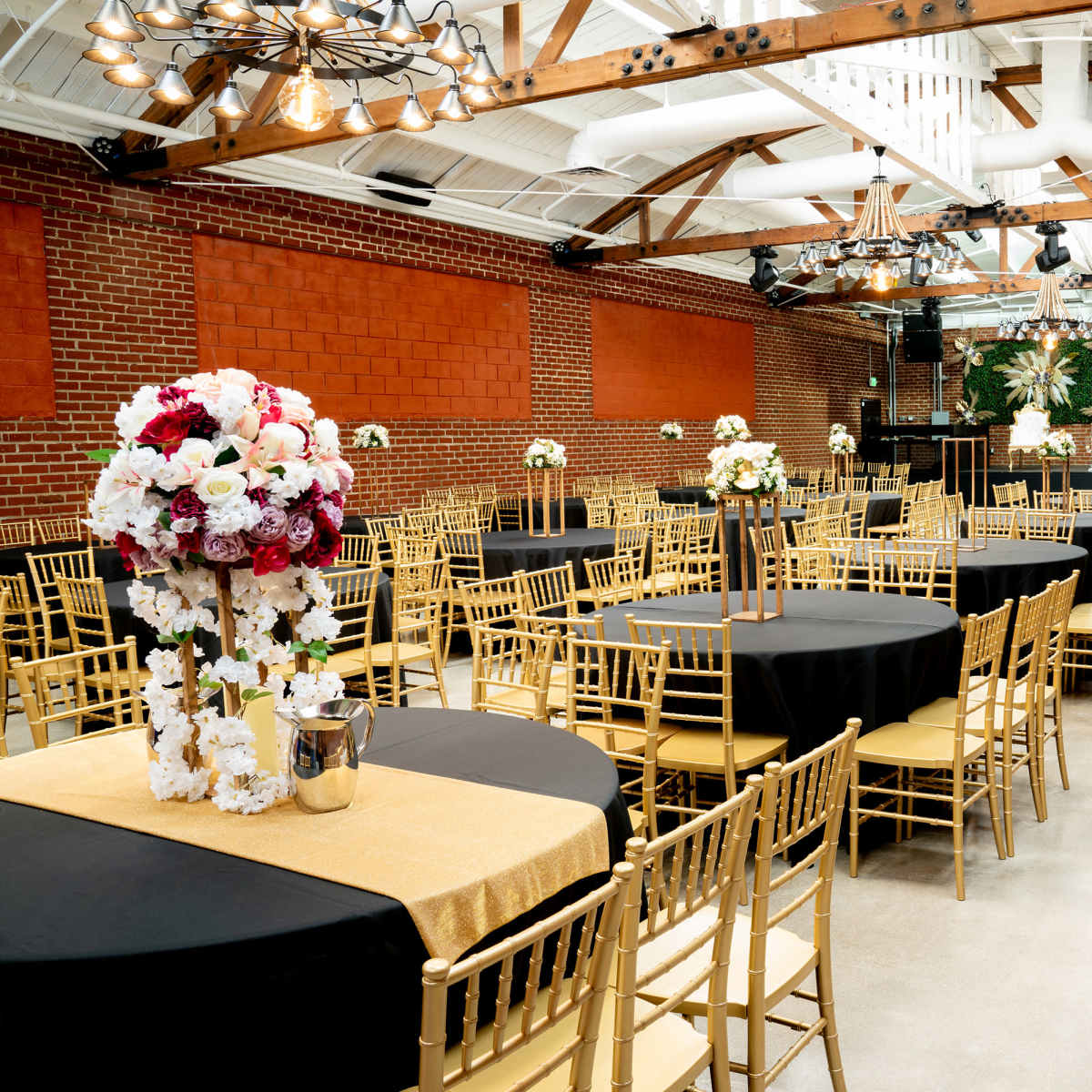 An elegantly arranged event space with rows of round tables covered in black and gold tablecloths, surrounded by gold chiavari chairs and decorated with floral centerpieces.