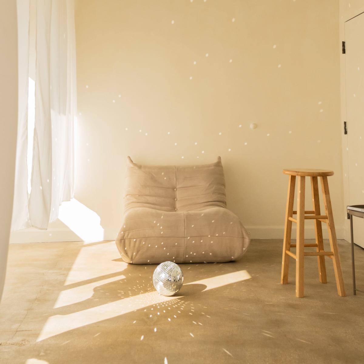 A quiet room features a beige floor chair and a wooden stool, with sunlight casting dots on the walls from a disco ball on the floor.