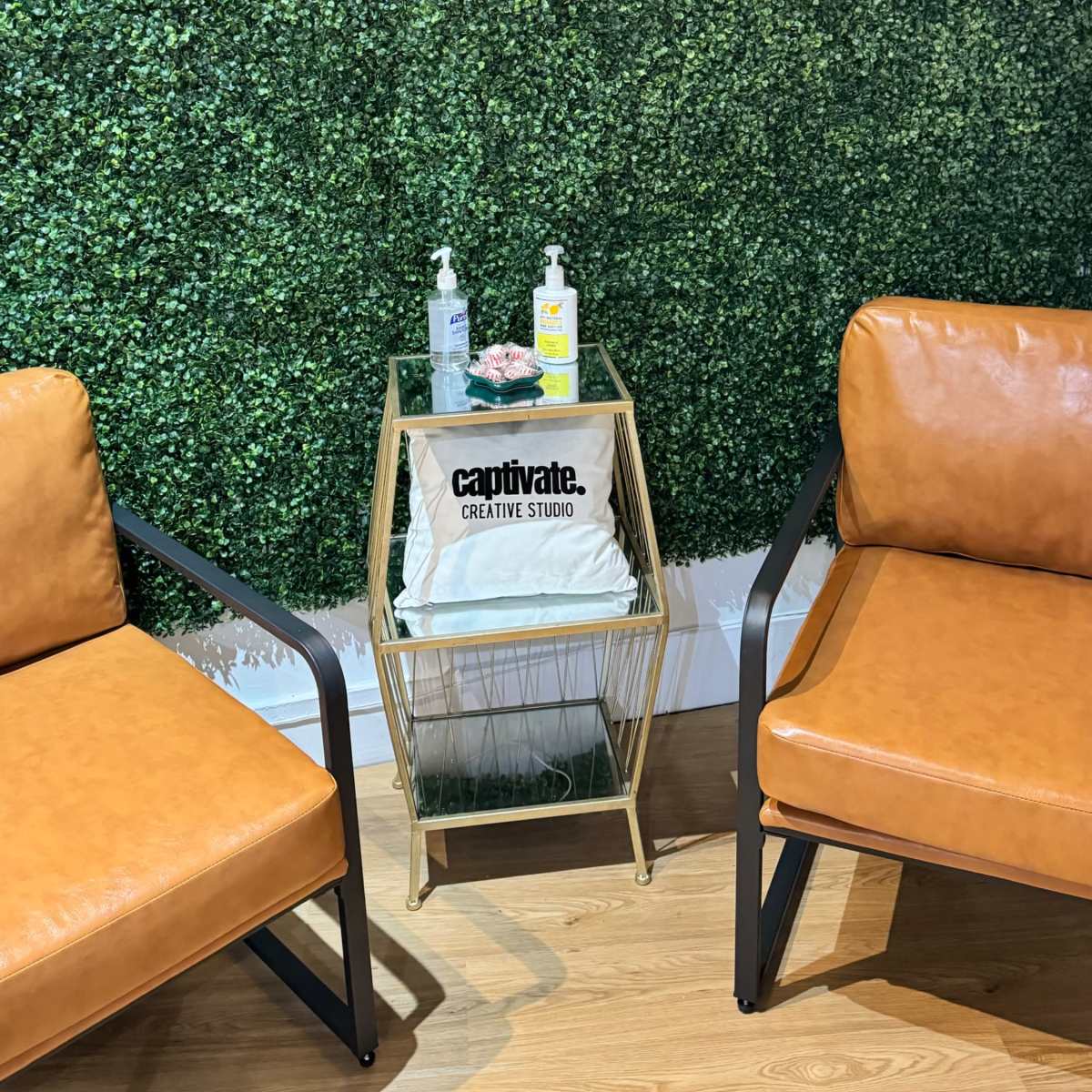 Two brown leather chairs facing a small gold side table, which holds hand sanitizers and a decorative item, against a backdrop of a green artificial hedge with a neon sign that reads "Pretty and Peaceful."