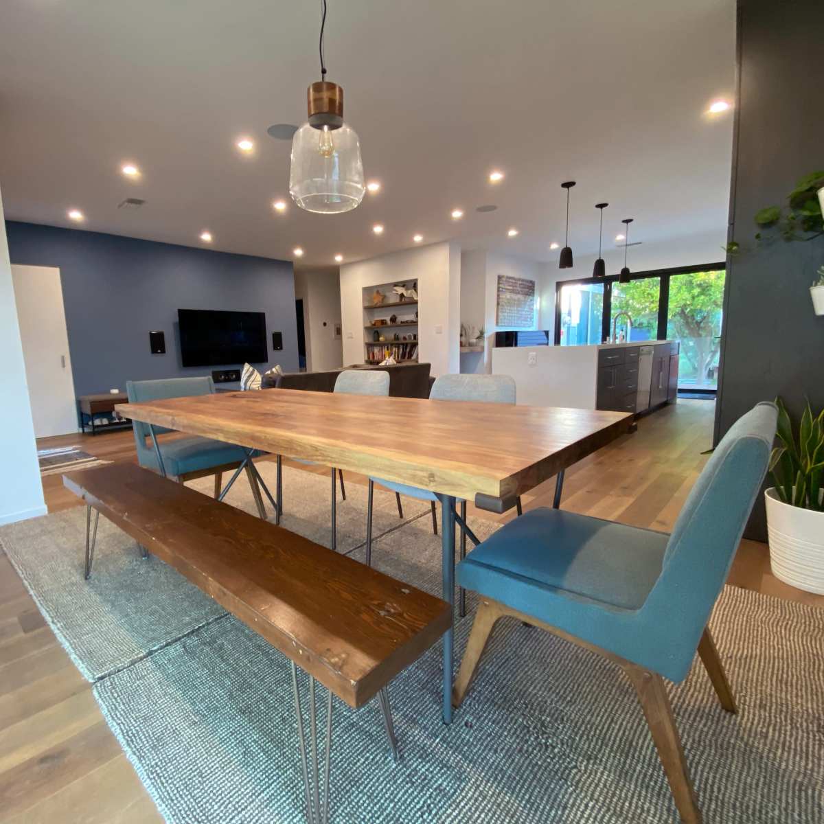 A modern dining room with a wooden table, blue chairs, and a bench, surrounded by large windows and contemporary decor.