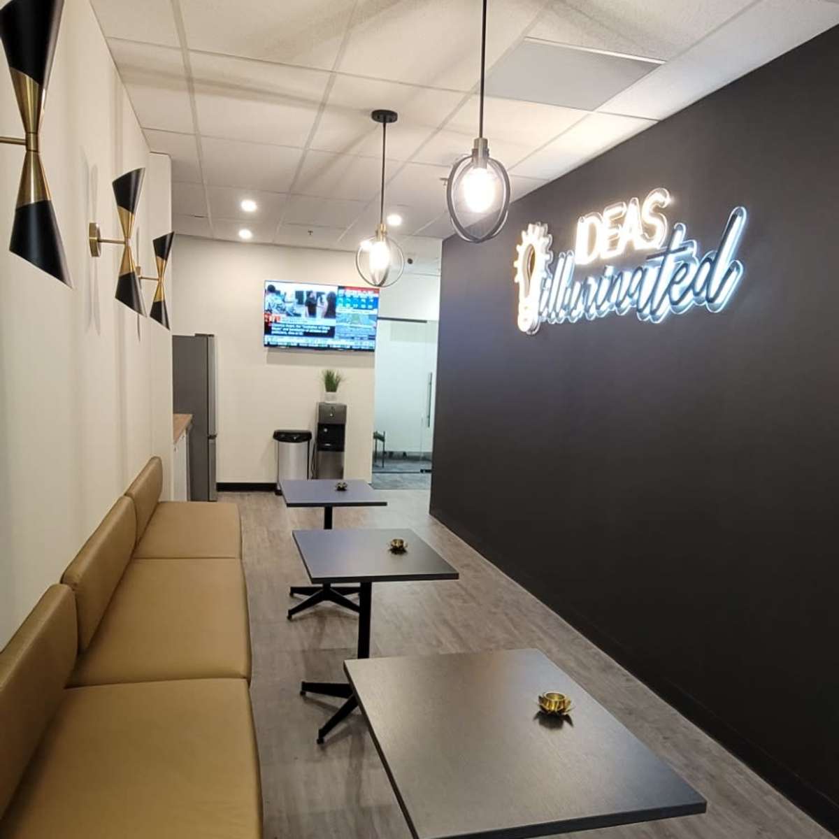 A modern workspace with a seating area featuring a tan bench and several black tables, illuminated by pendant lights, and a wall displaying the phrase "Ideas Unlimited" in neon.