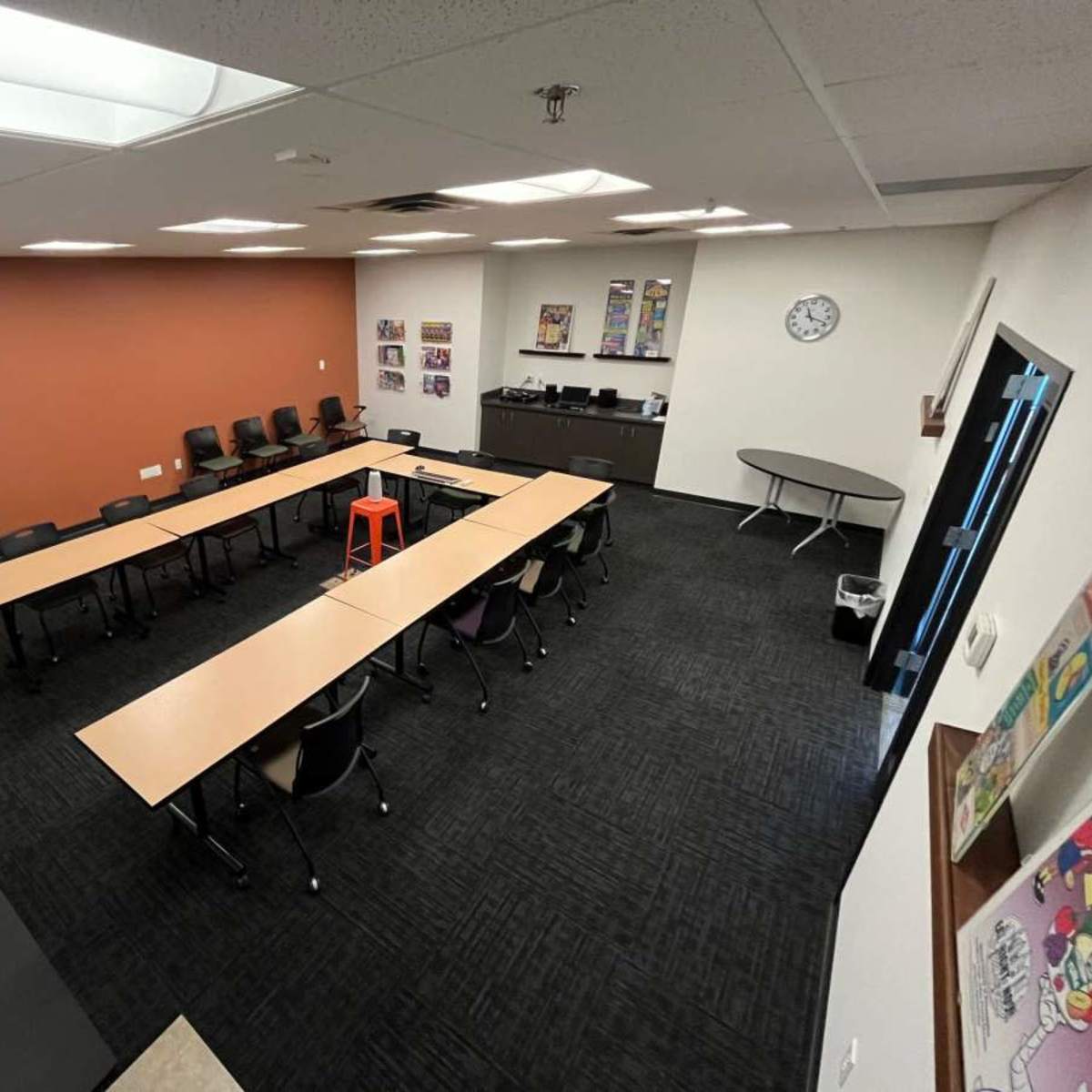 A classroom with several tables arranged in a u-shape, surrounded by chairs, and featuring an orange stool in the center.
