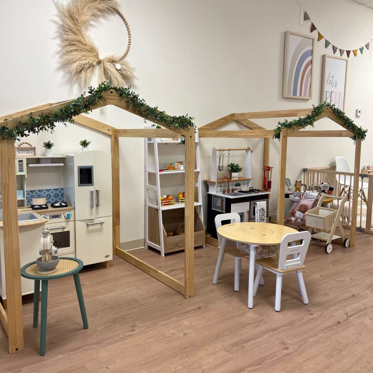 A children's play area featuring two playhouses with wooden frames, a small table with chairs, and various toys and kitchen sets displayed on shelves.