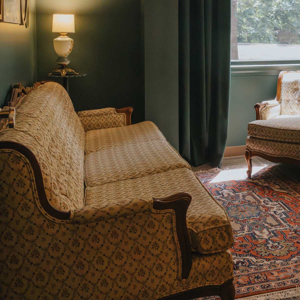 A cozy sitting area featuring a patterned sofa, an armchair, a side table with a lamp, and a colorful area rug.