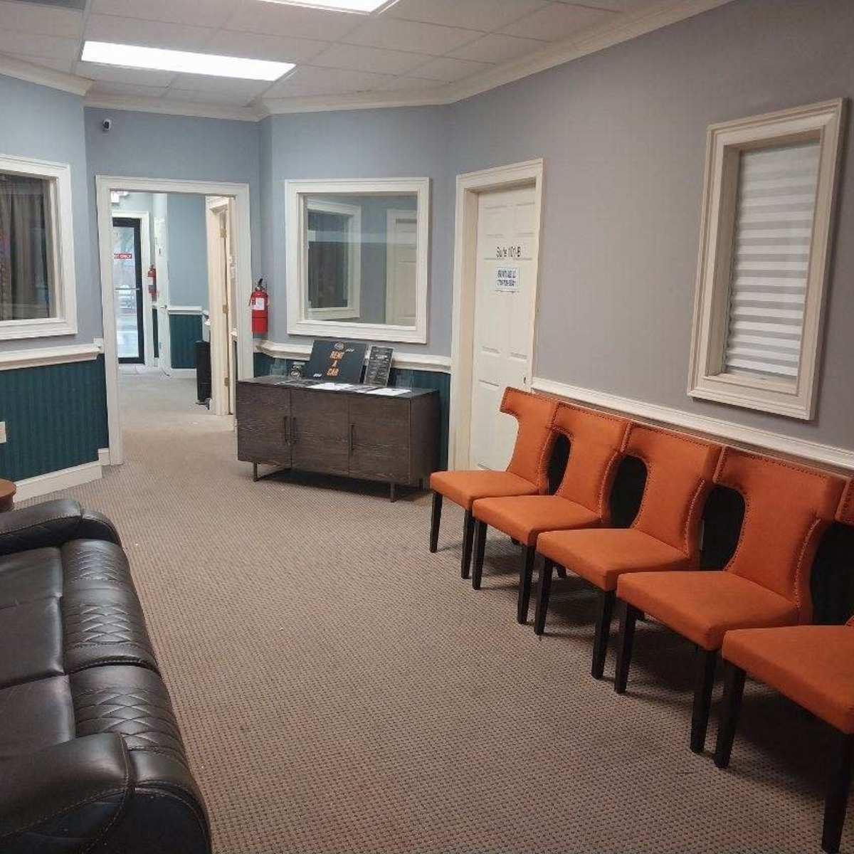 A waiting room with a black leather couch and a row of orange chairs against the wall, framed by light gray walls and two doors leading to other rooms.