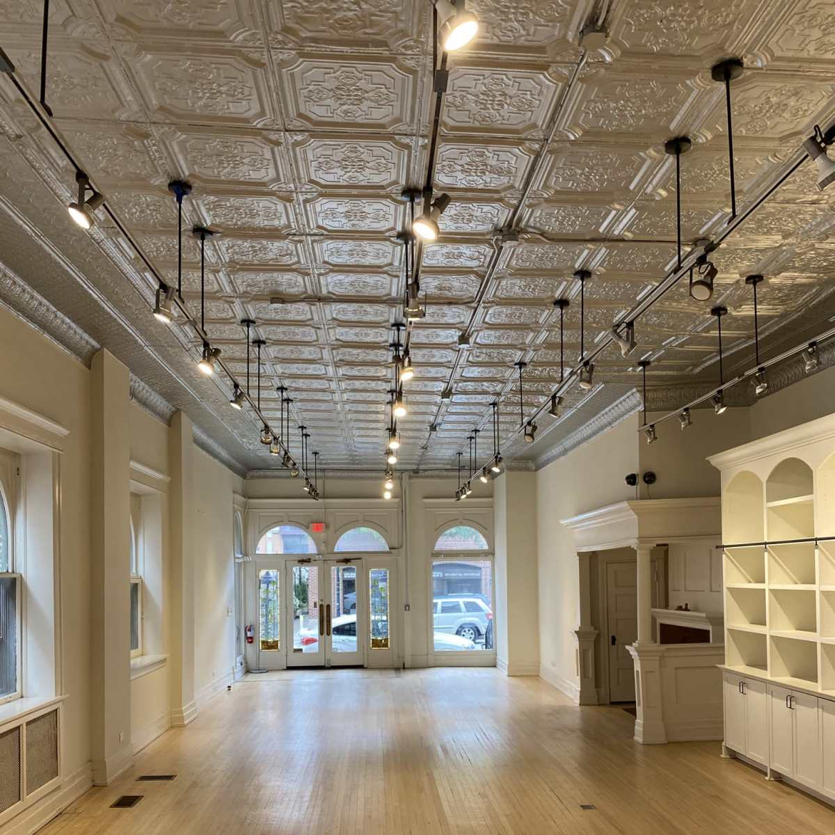 An empty retail space with a wooden floor, decorative ceiling tiles, and large front windows.