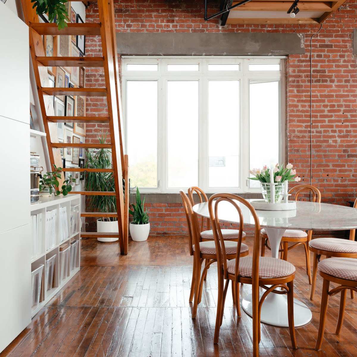 A bright, airy dining area with a round table surrounded by bentwood chairs, a staircase leading to a loft, and exposed brick walls.