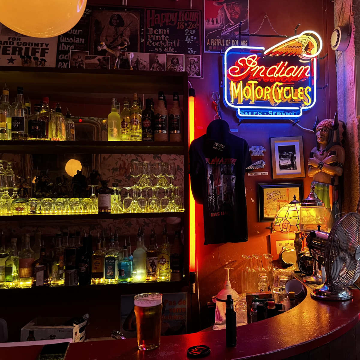 The bar features a neon sign reading "Indian Motorcycles" alongside shelves filled with various bottles and a fan in the background.