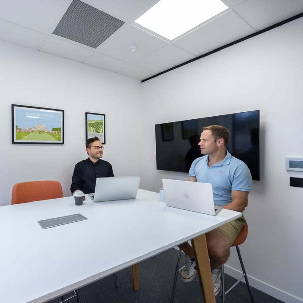 Two men are seated at a table in a bright meeting room, discussing work while using laptops.