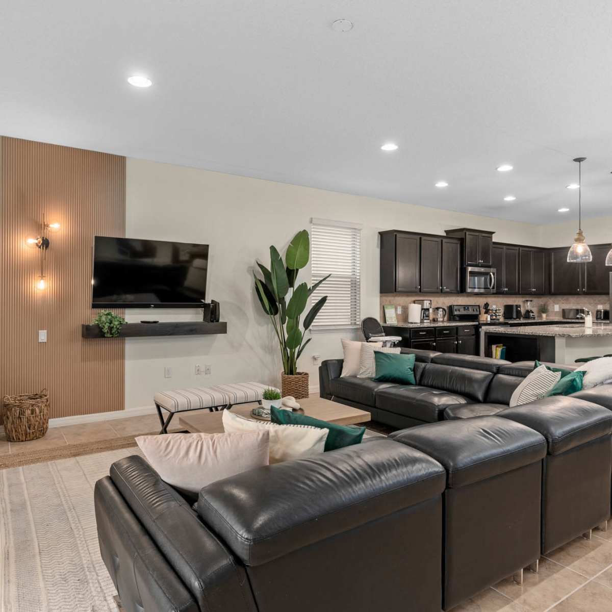 A modern living room with a dark leather sectional sofa, a wooden accent wall, and a kitchen area in the background featuring dark cabinetry and pendant lighting.