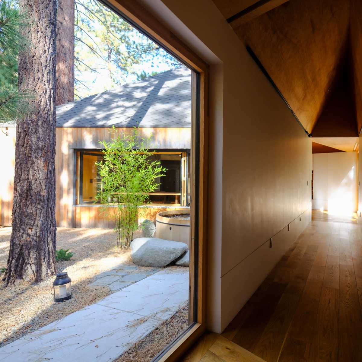A narrow hallway with wooden flooring leads to a bright open space, framed by a large window showing a tranquil outdoor area with trees and rocks.