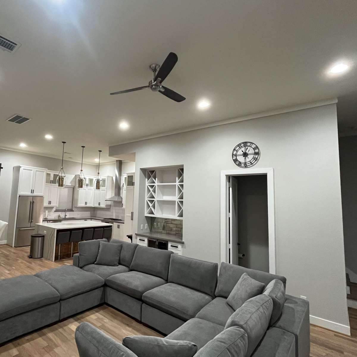 A modern living room featuring a large gray sectional sofa, hardwood flooring, and an open-concept layout that connects to a dining area and kitchen.