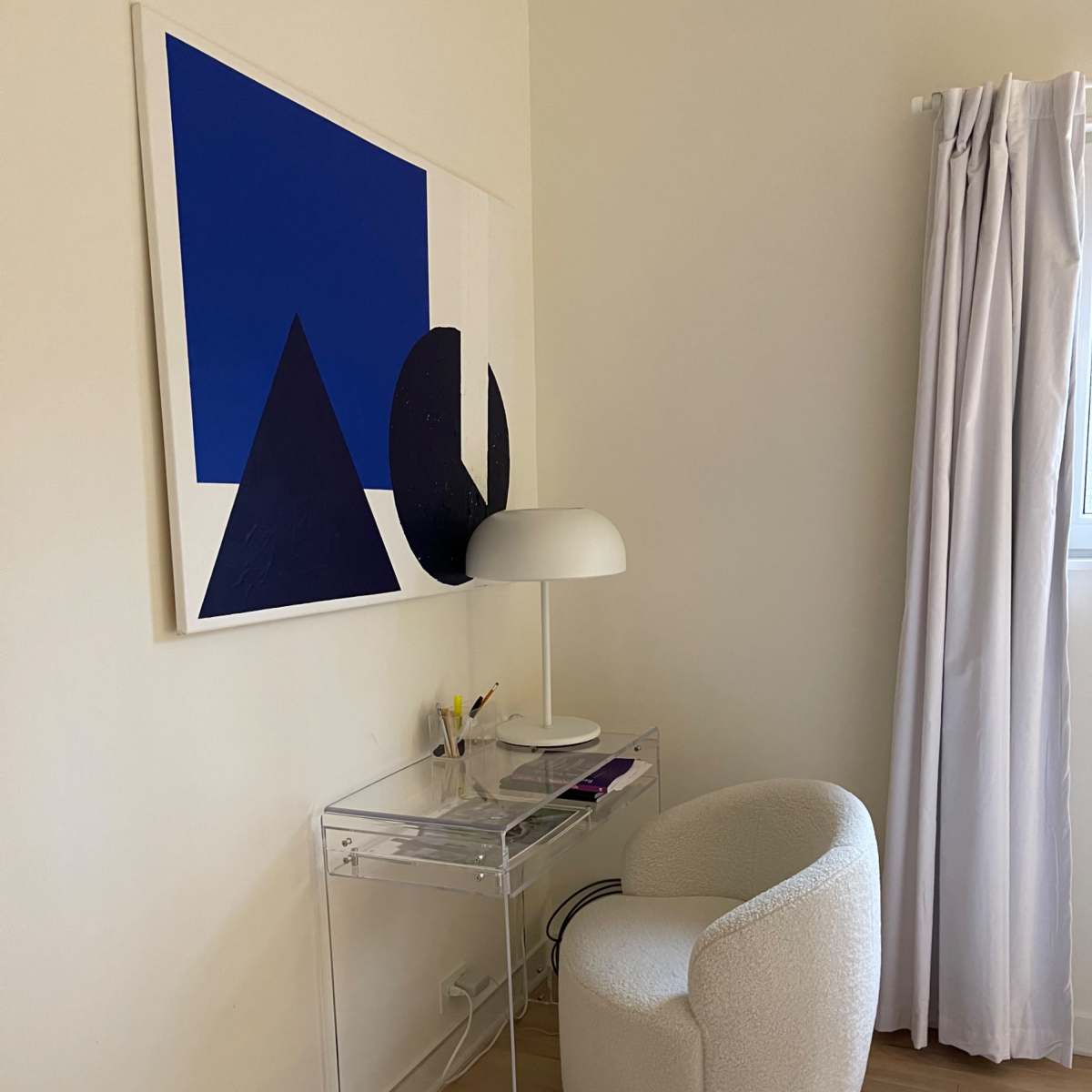 A clear acrylic desk with a white lamp and art piece hangs on the wall beside a round, upholstered chair.