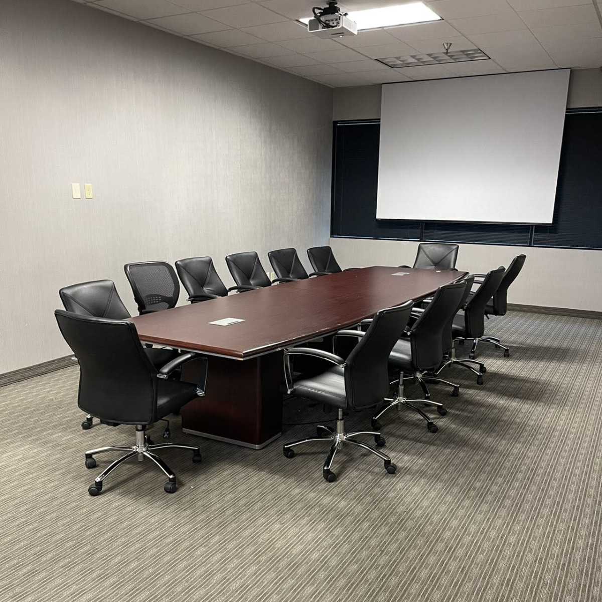 A long conference table is surrounded by office chairs in a sparsely furnished meeting room.
