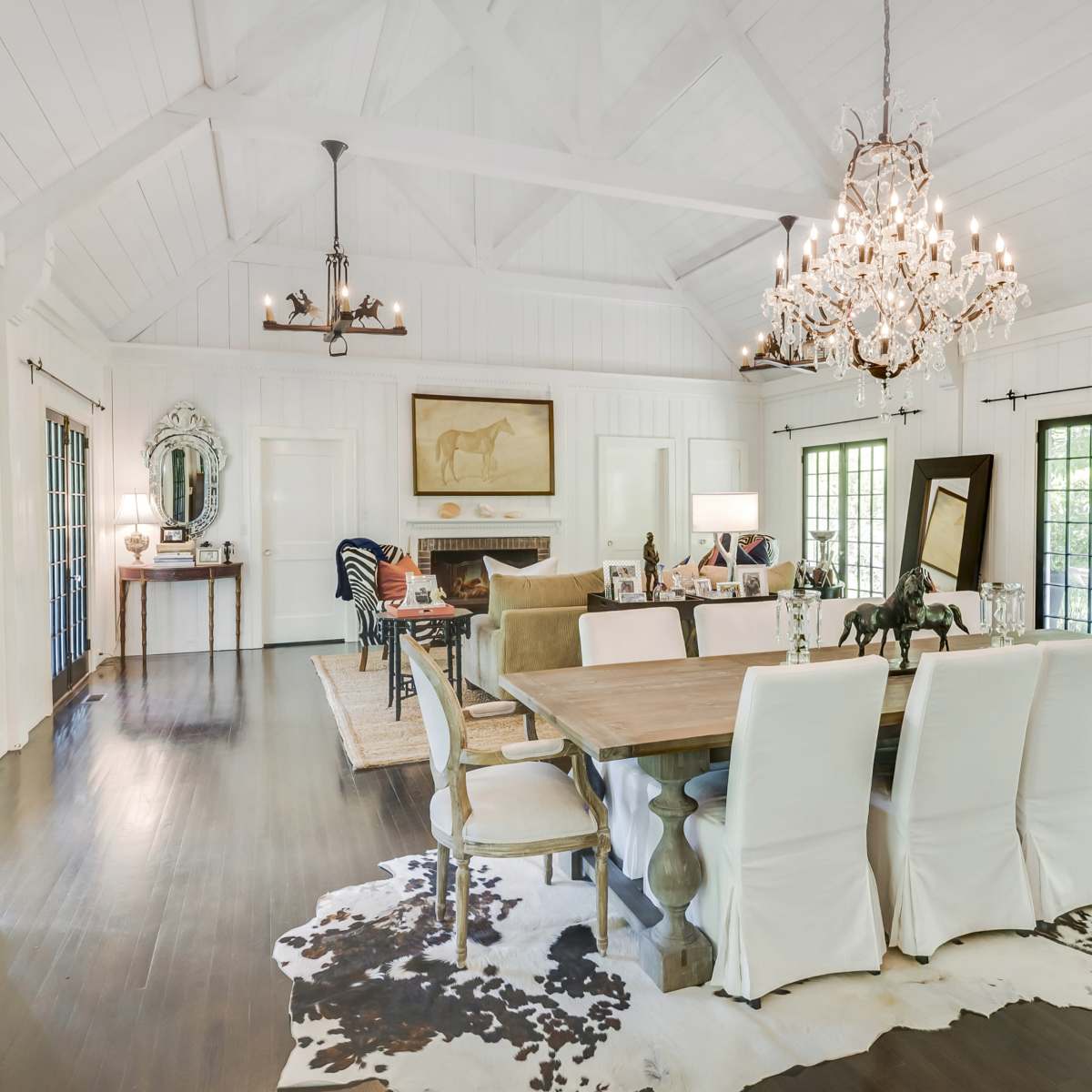 A spacious dining room featuring a large wooden table surrounded by white upholstered chairs, with a chandelier overhead and a mix of decorative elements and artwork on display.