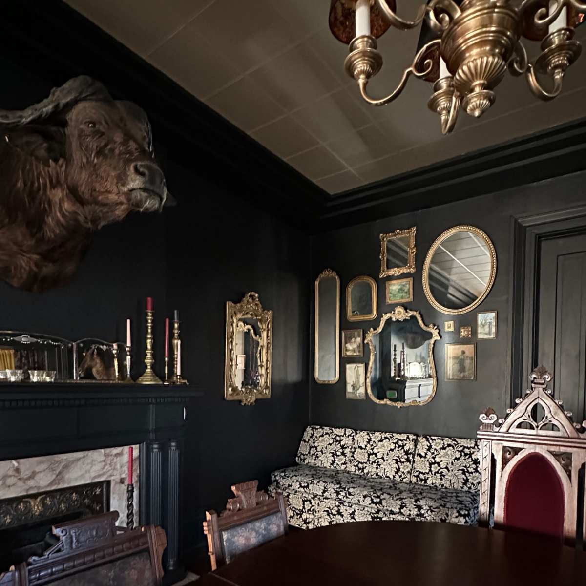 The dimly lit dining room features a dark wall, a mounted animal head above an ornate fireplace, a patterned sofa, and an arrangement of framed mirrors and artwork.