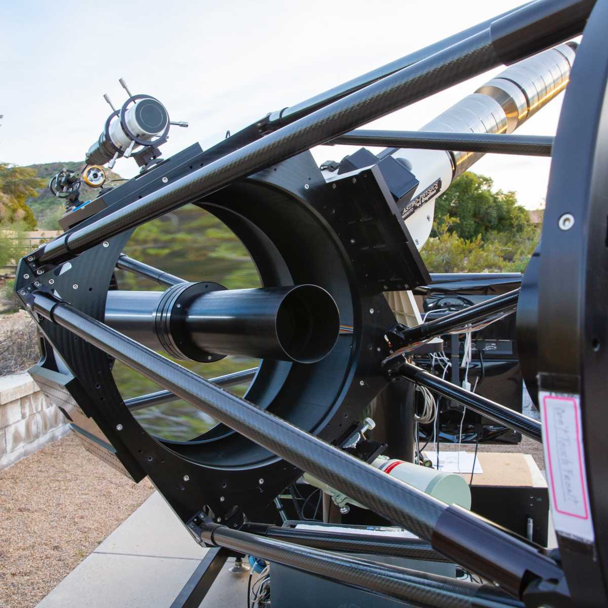 A large telescope with a black carbon fiber frame is positioned outdoors, pointing towards the sky.