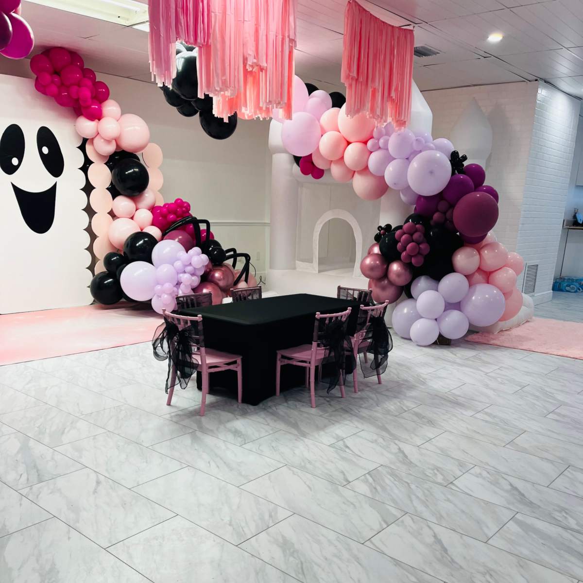 A decorated event space featuring a black table with pink chairs surrounded by colorful balloon arrangements and a wall with a ghost design.