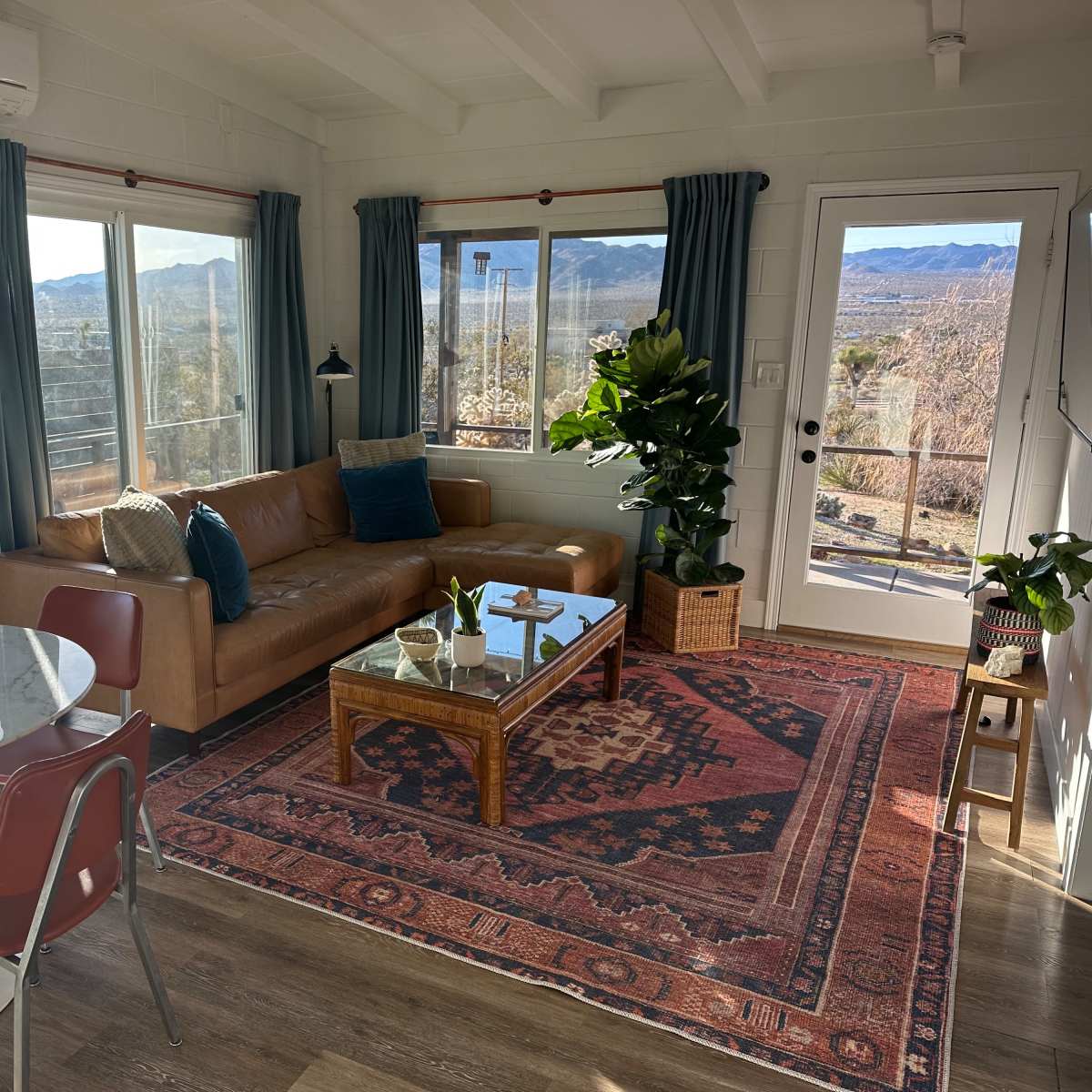 A living area featuring a brown leather couch, a glass coffee table on a patterned rug, a dining table with red chairs, large windows with mountain views, and potted plants.