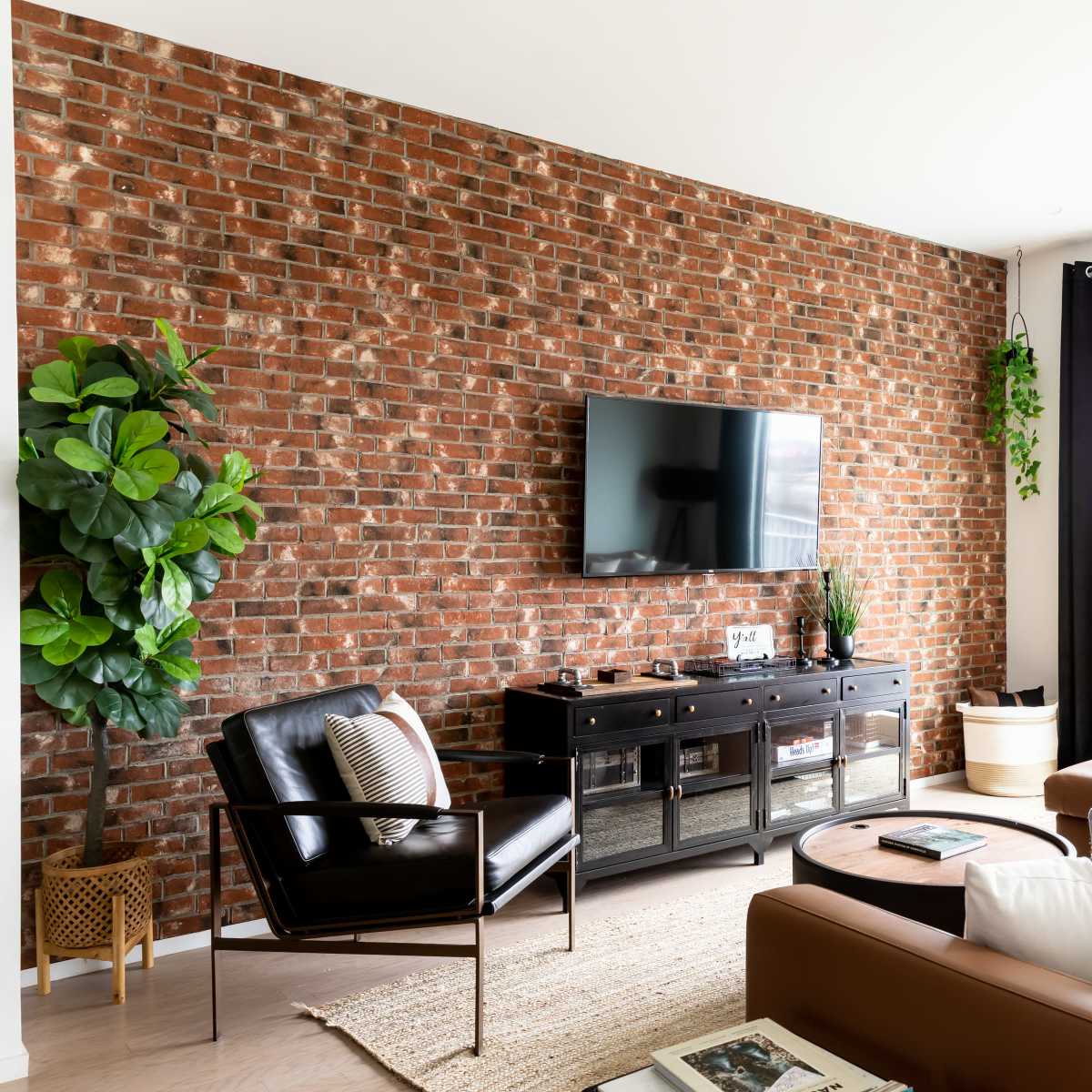 A modern living room features a brick accent wall, a large TV mounted above a dark sideboard, and a sectional sofa near a window with a view.