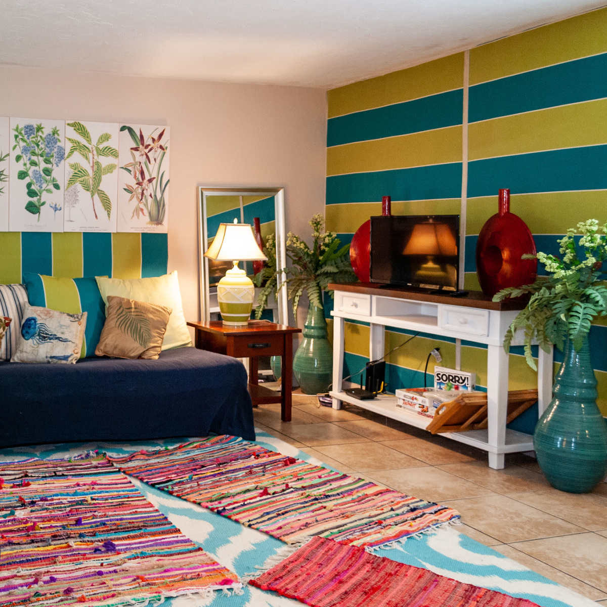 A brightly decorated living room with a blue couch, colorful throw pillows, and striped walls, featuring a coffee table and lamps.
