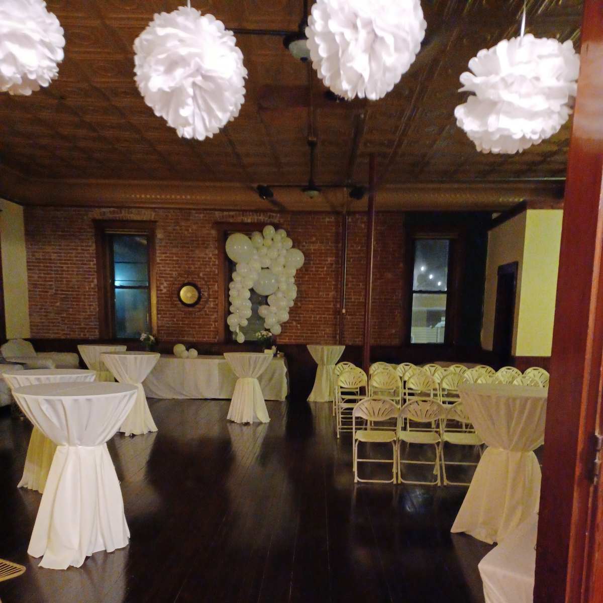 An empty event space with white tablecloths draped over several round tables and white paper decorations hanging from the ceiling.