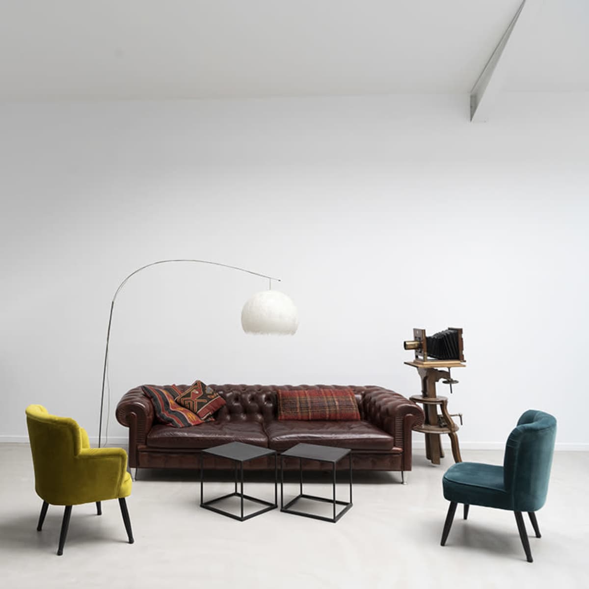 A living room with a brown leather chesterfield sofa, two colorful chairs, a floor lamp, and a wooden shelf against a white wall.