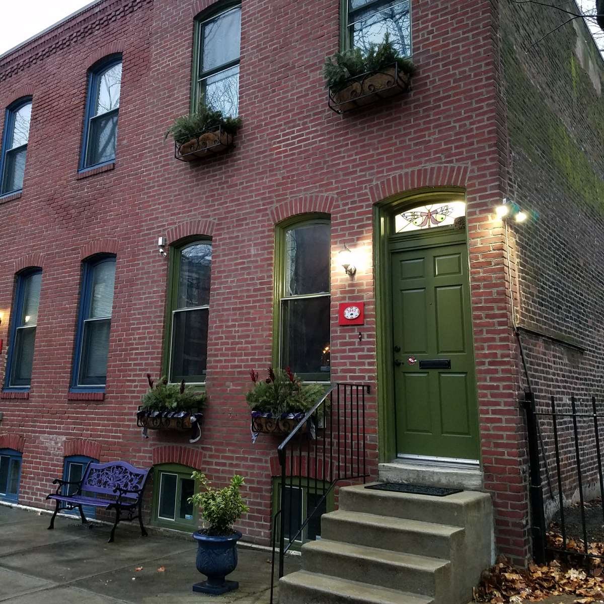 A brick townhouse with green door and window frames, featuring potted plants on the windowsills and a small staircase leading to the entrance.