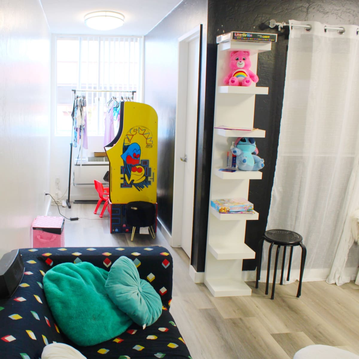 A narrow hallway with a colorful couch, a vintage arcade game cabinet, a clothing rack, and a shelving unit containing toys and books.