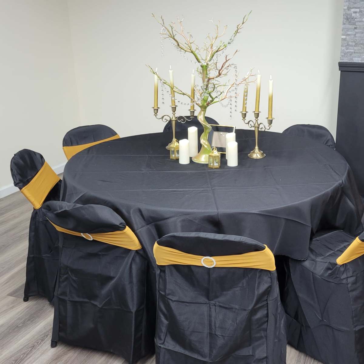 A round table covered with a black tablecloth and adorned with gold accents is surrounded by chairs dressed in black covers with yellow sashes.